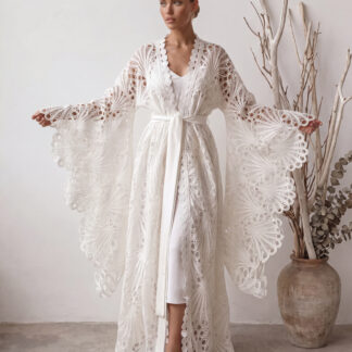 Front view of a bohemian white lace kimono with wide flared sleeves and a peacock feather cutout pattern. Model with a sleek bun stands in a minimalist studio, arms outstretched to showcase the flowy, floor-length design.